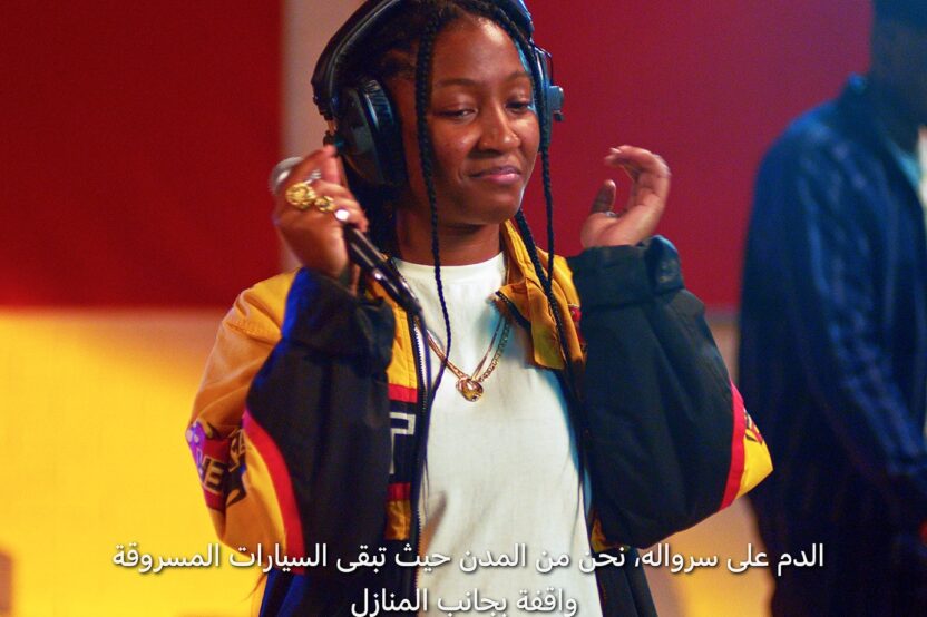 Jeune femme portant un casque audio, esquissant un sourire, avec un sous-titre en arabe en bas de l’image.