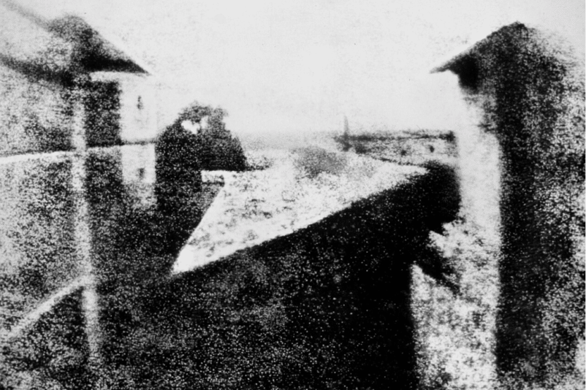 Photographie ancienne en noir et blanc, très granuleuse, montrant une table rectangulaire recouverte d’une nappe à motifs, entourée de silhouettes humaines devant des bâtiments.