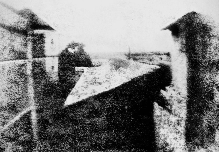 Photographie ancienne en noir et blanc, très granuleuse, montrant une table rectangulaire recouverte d’une nappe à motifs, entourée de silhouettes humaines devant des bâtiments.